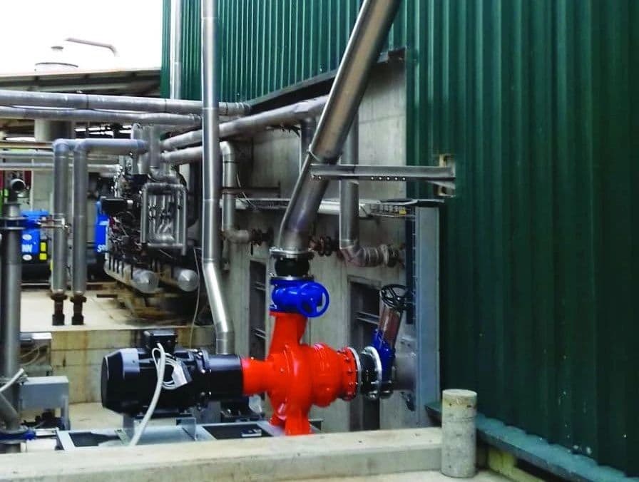 A robust CRI-MAN industrial pump, painted bright orange, installed on a concrete platform next to a large, green
corrugated metal storage tank. The pump is connected with heavy-duty pipes and valves, showcasing a clean and professional installation for fluid transfer.